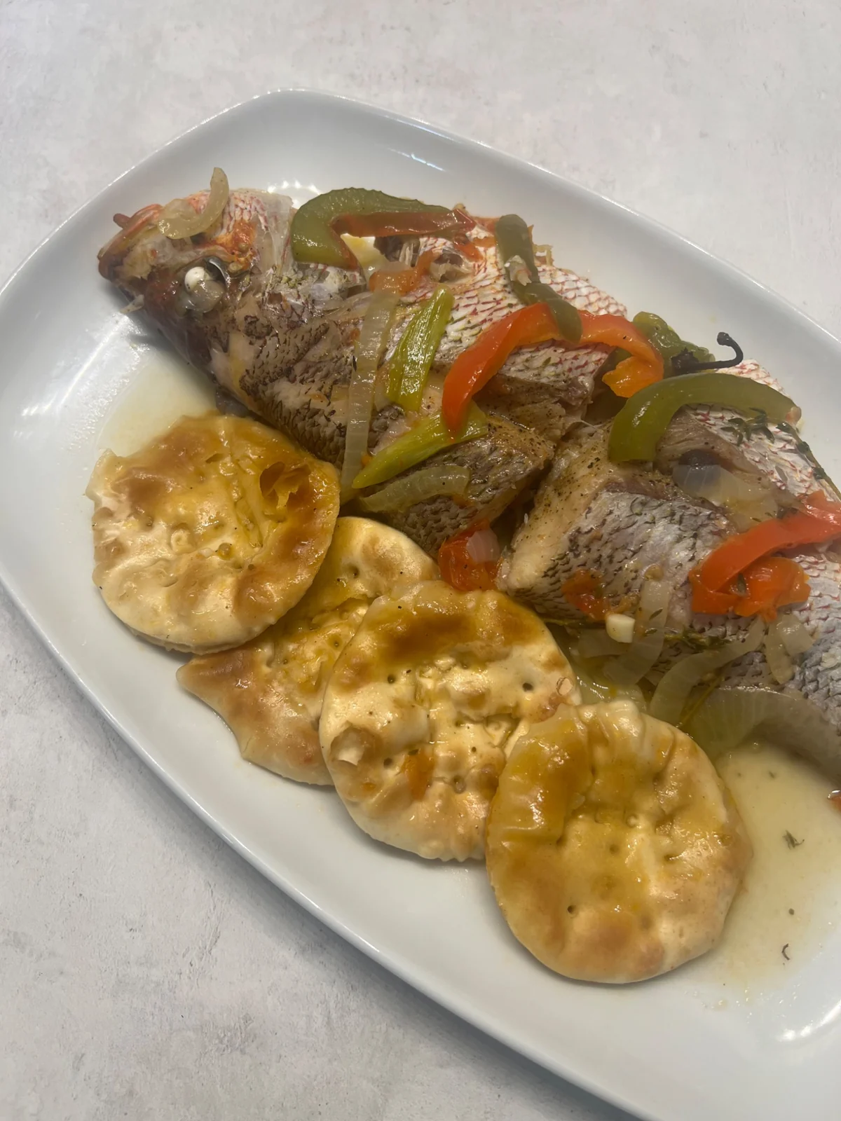 Jamaican steamed snapper with vegetables and whole gravy-soaked Excelsior crackers on a white platter